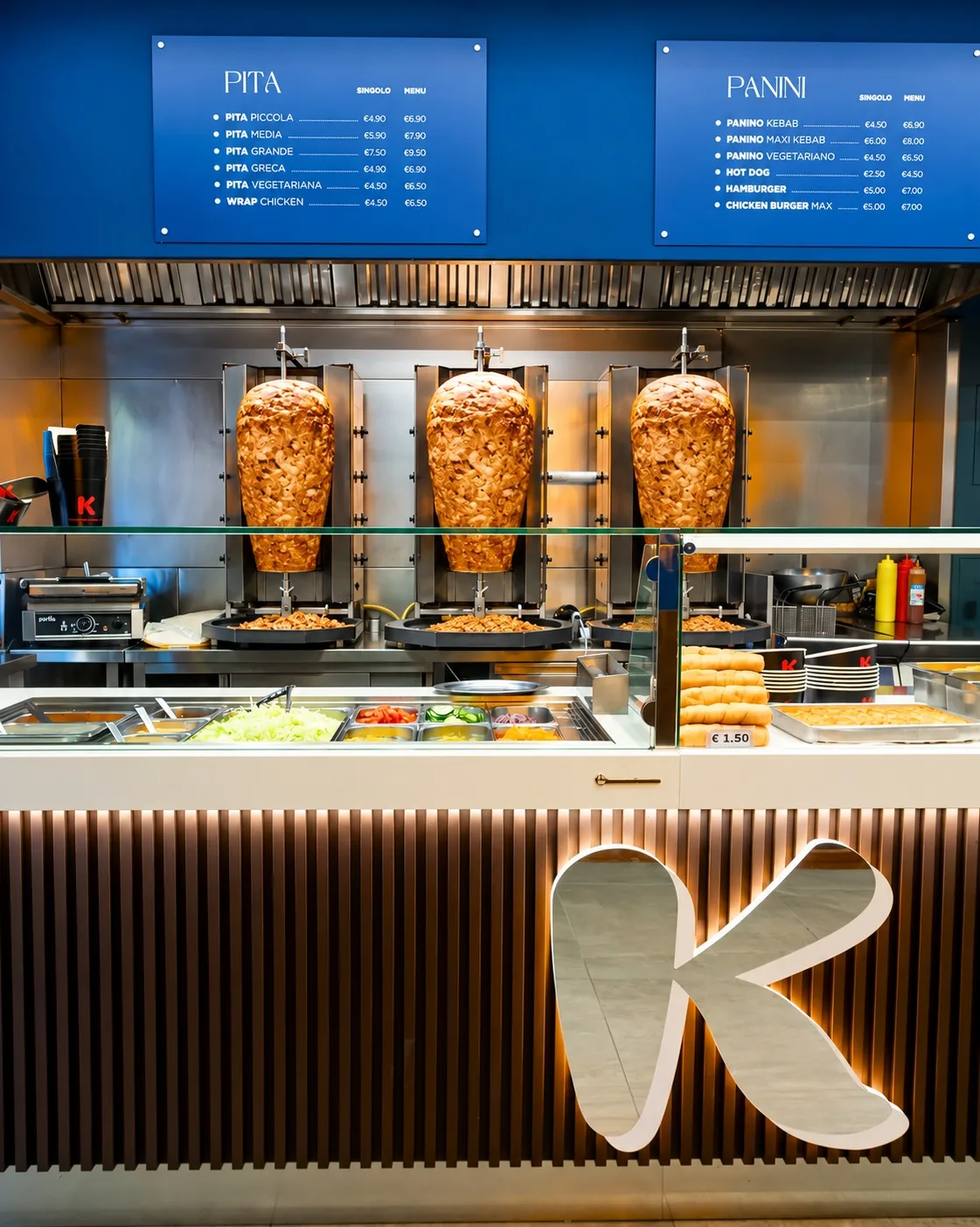 Un bancone Kebabberia: tre spiedi di döner, vaschette di insalata fresca e la grande K